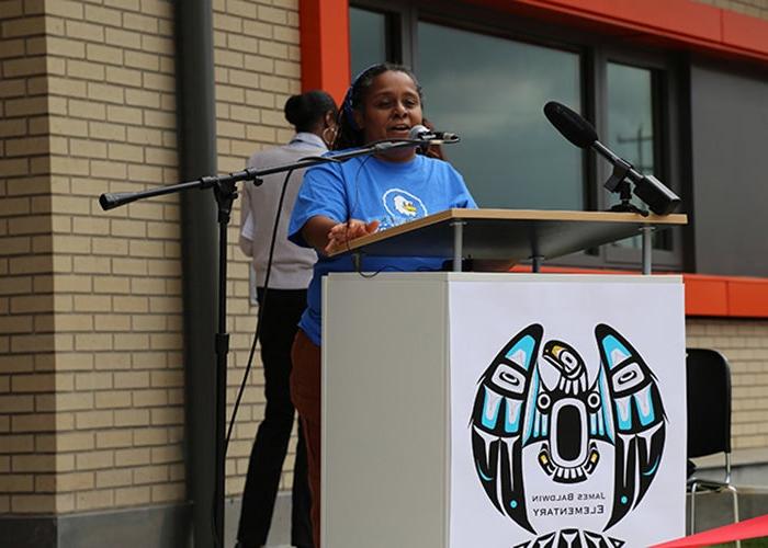 a woman stands at a podium with microphones attached. a light colored brick wall and a window with an orange frame are behind her