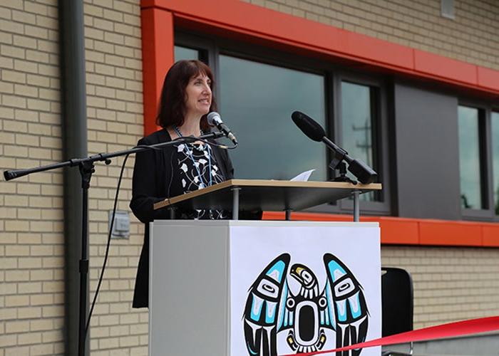 a woman stands at a podium with a microphone attached. a light colored brick wall and a window with an orange frame are behind her